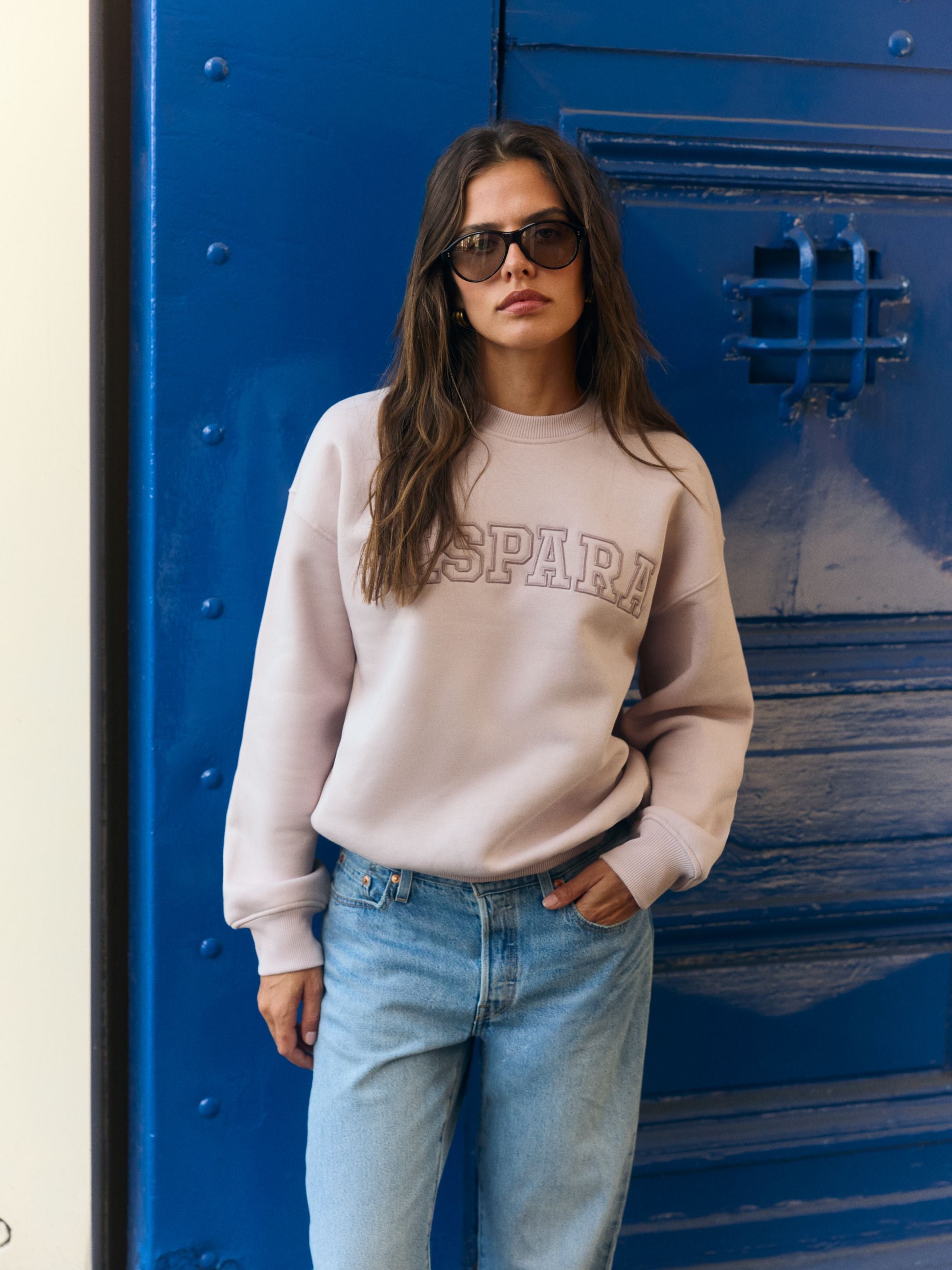 Woman wearing a dusty pink CASPARA logo crewneck sweatshirt and sunglasses against a blue door.